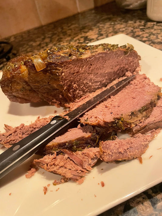 Query successful A cooked and sliced pot roast, seasoned with herbs, on a white platter with a carving knife.