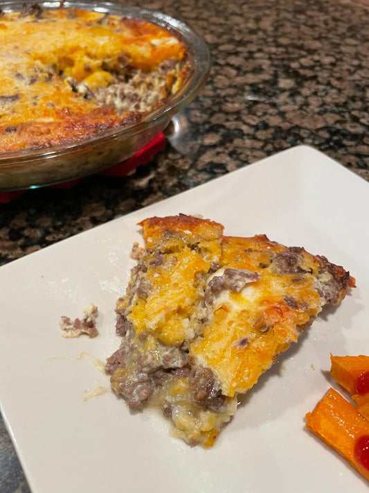 A slice of golden-brown cheeseburger casserole on a white plate with carrots, and the full casserole dish in the background.