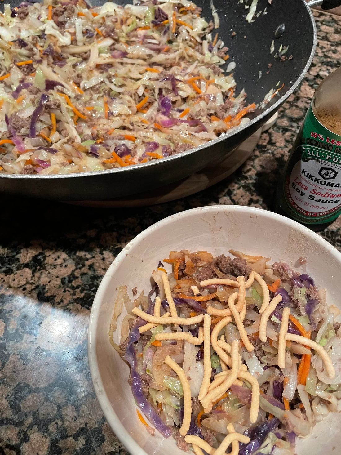 White bowl of "Egg Roll in a Bowl" topped with crispy noodles, with a pan of the mixture and soy sauce bottle in the background.