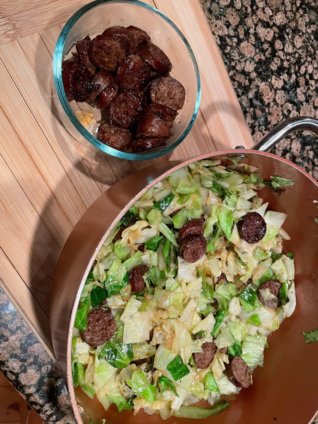 A large copper pot filled with sautéed cabbage and sliced beef sausage, with an extra bowl of sausage on a wooden board.