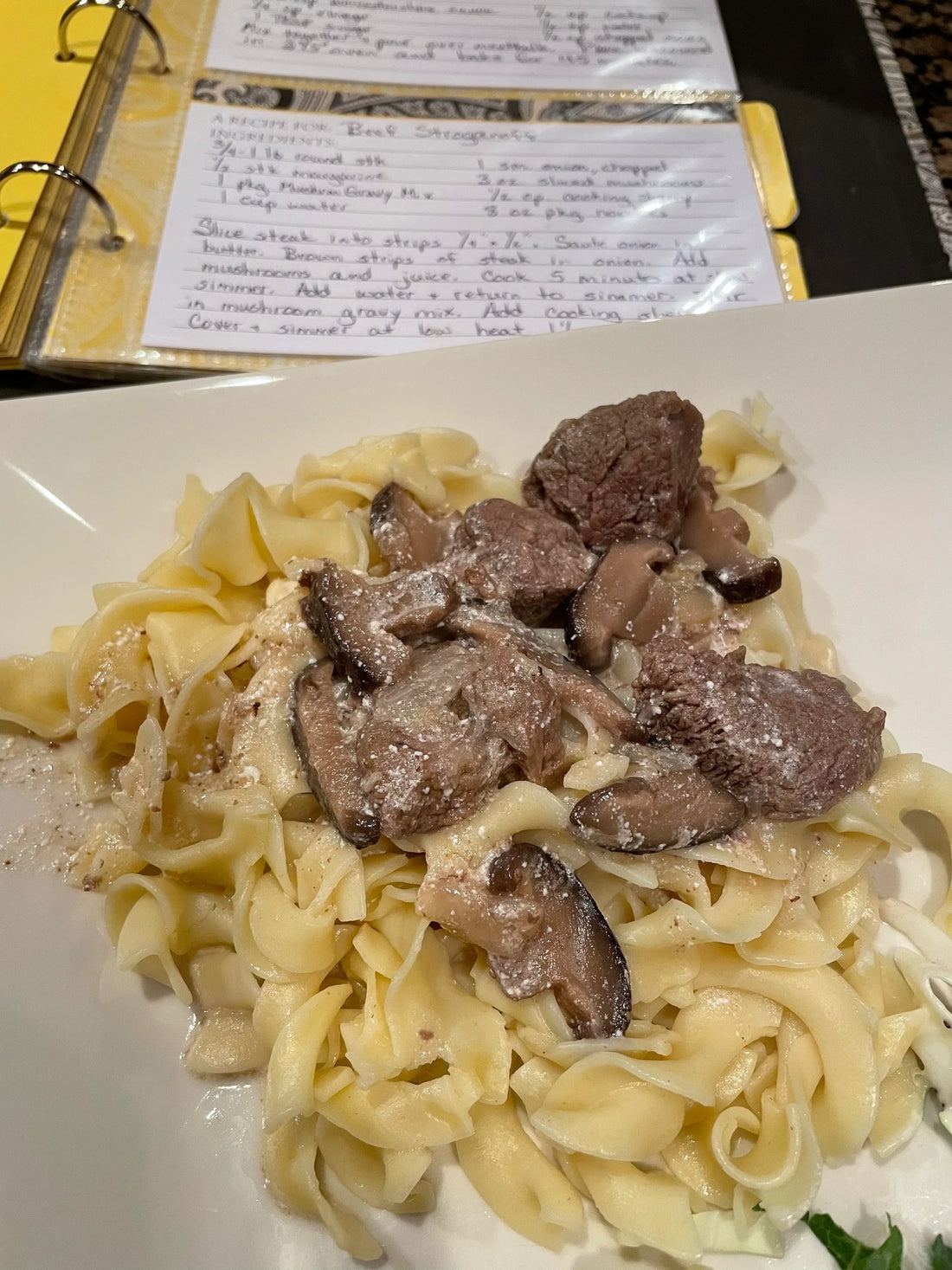 Beef Stroganoff on a white plate with egg noodles, beef, mushrooms, and creamy sauce, with a handwritten recipe visible in the background.